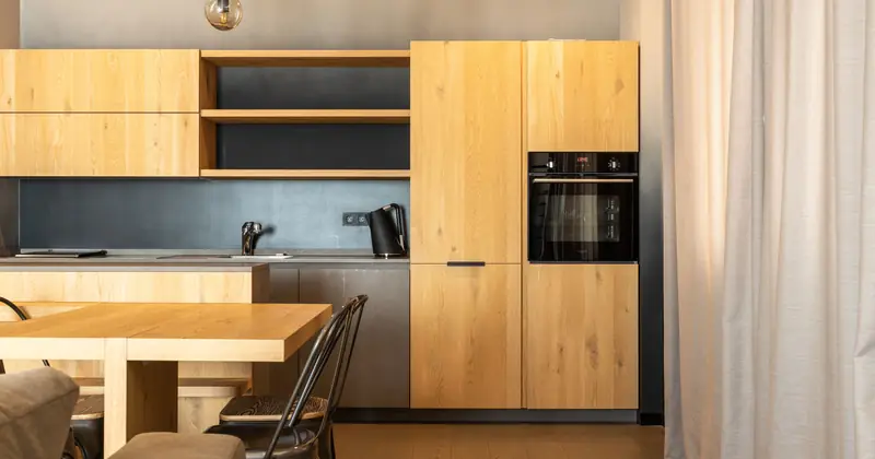 Wooden table with chairs placed in stylish kitchen with cupboards and modern appliances in apartment with curtains on window and couch