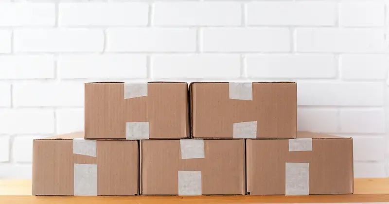 A neat stack of cardboard boxes taped and placed on a wooden shelf against a white brick wall.