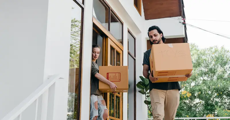 Young movers carrying cardboard boxes out of a house on a summer moving day.